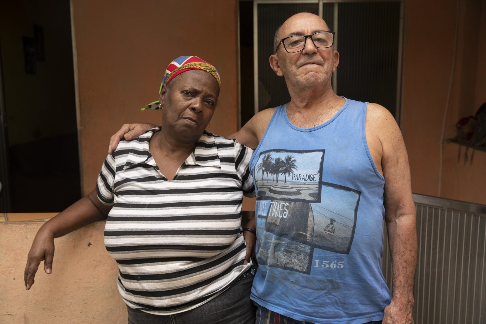 Vera Lúcia e José Luiz, avós de Diego, jovem morto pelo exército em 2018. À esquerda, Vera Lúcia, uma mulher negra na faixa dos 60 anos. Ela veste uma camiseta branca com listras cinzas e pretas, além de uma bandana estampada na cabeça. À direita está José, um homem branco de regata azul estampada com cartões postais do Rio de Janeiro. Os dois estão abraçados na entrada de casa.