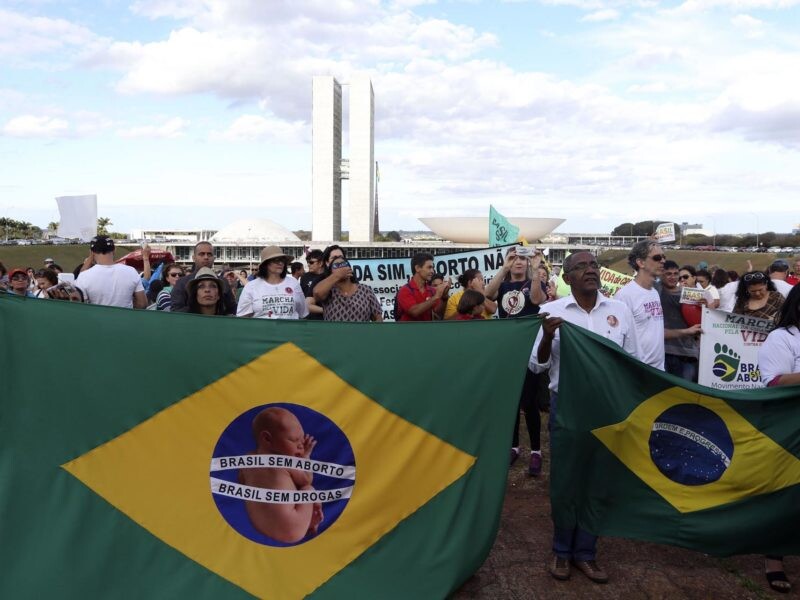 Cerca de 100 manifestantes na Esplanada dos Ministérios, em Brasília. Mais a frente da imagem, pessoas seguram duas bandeiras do Brasil e em uma está escrito: Brasil sem aborto, Brasil sem drogas