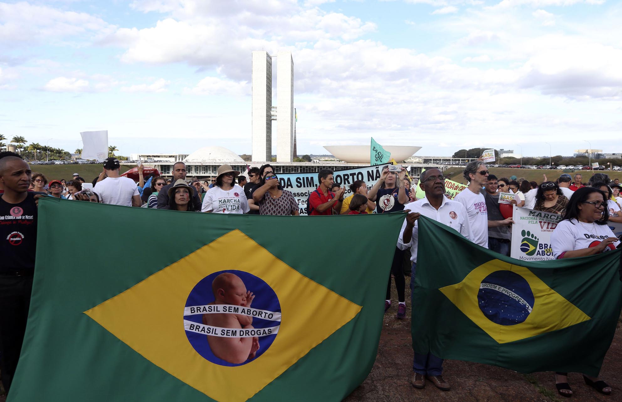 Cerca de 100 manifestantes na Esplanada dos Ministérios, em Brasília. Mais a frente da imagem, pessoas seguram duas bandeiras do Brasil e em uma está escrito: Brasil sem aborto, Brasil sem drogas