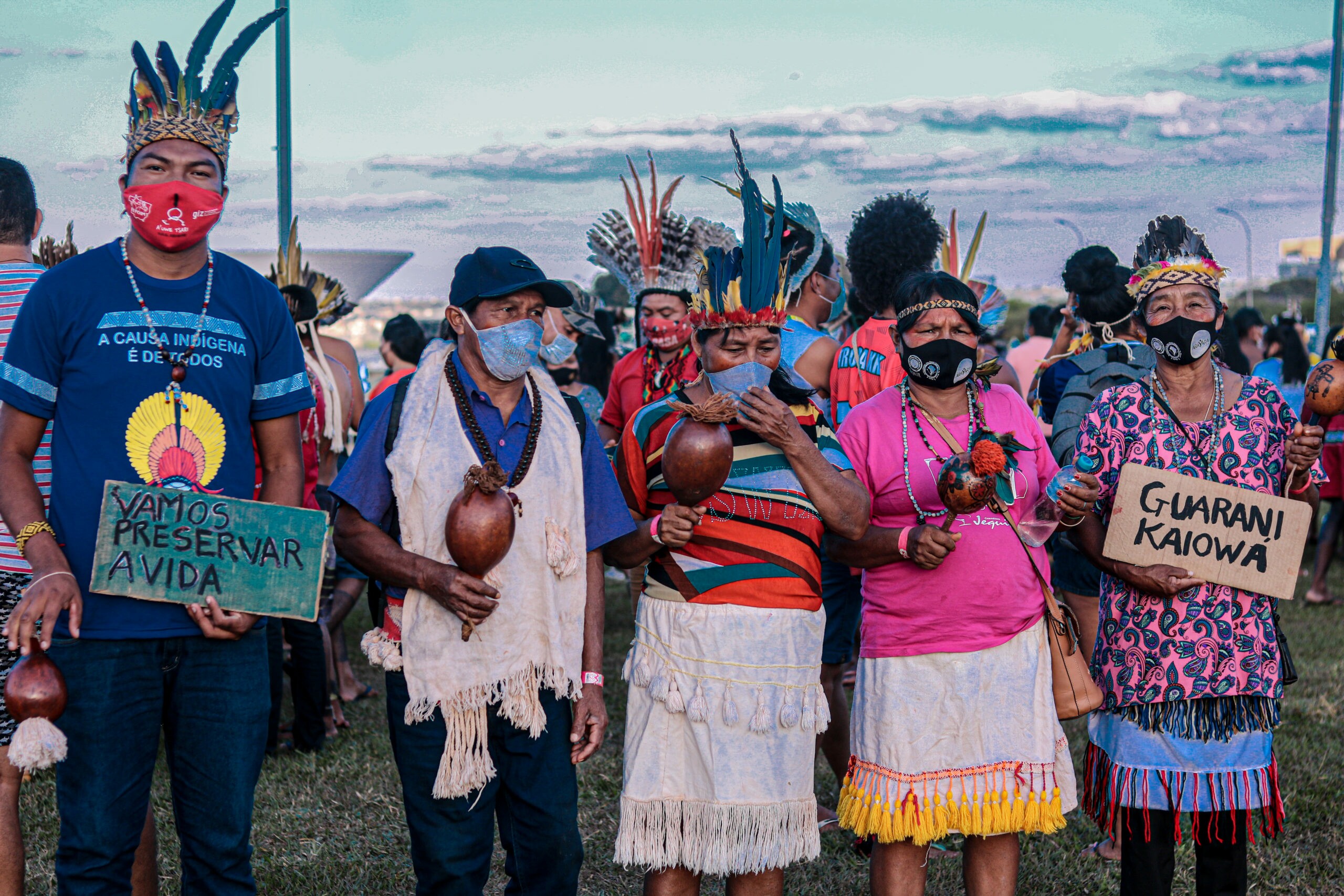 Indígenas vestidos com adereços tradicionais carregam placas com os dizeres 
