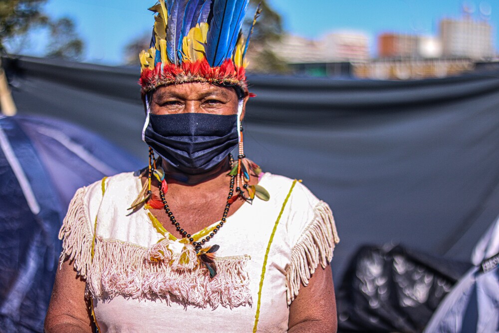 Mulher indígena posa de frente para a câmera, com adereços tradicionais na cabeça e bandeira preta ao fundo.