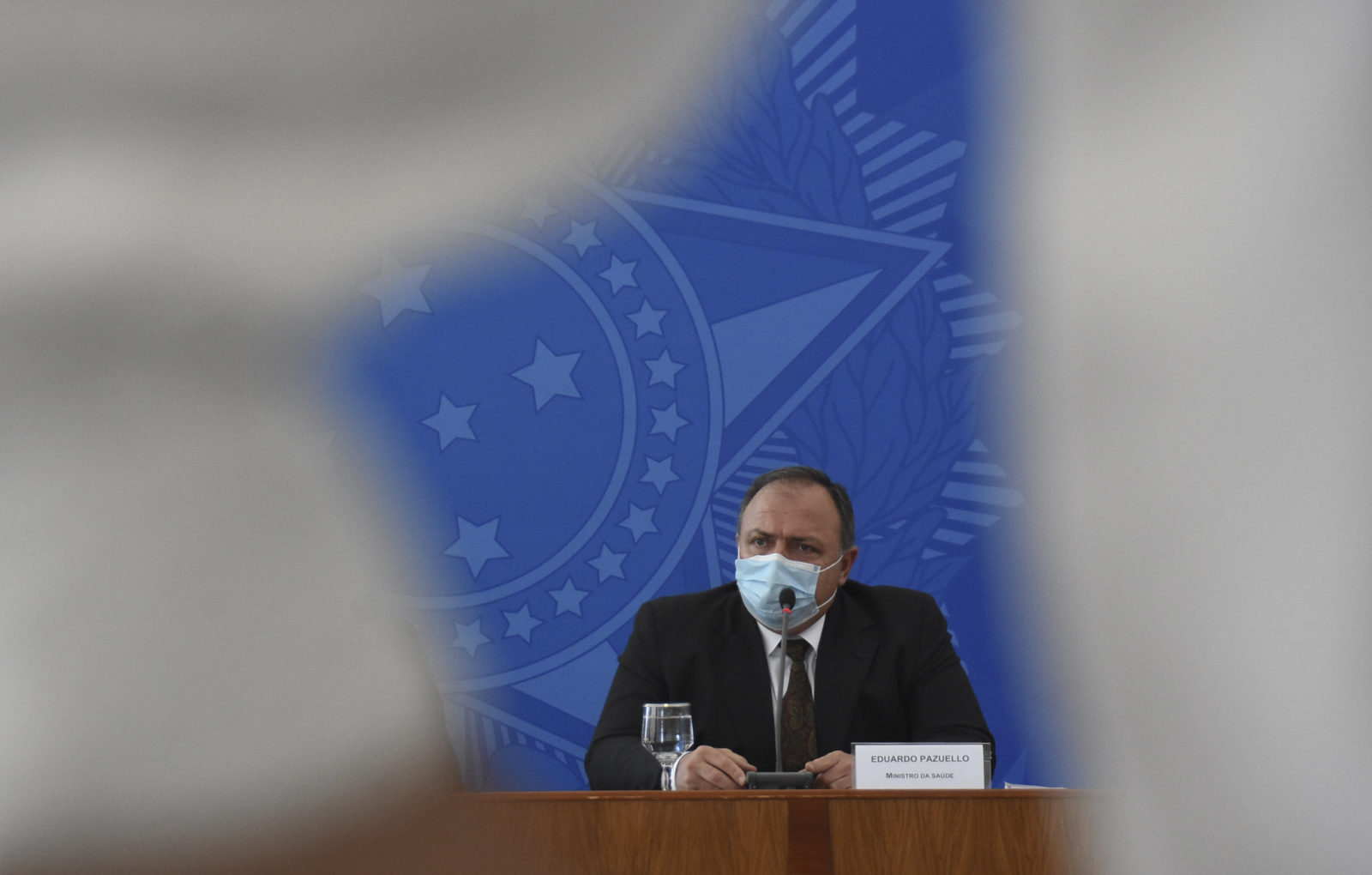 Pazuello, homem branco com cabelo curto, posa sentado de máscara descartável e terno preto, com o símbolo do Brasil atrás