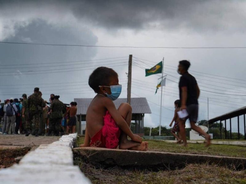 Criança indígena à beira da estrada com exército e bandeira do Brasil ao fundo