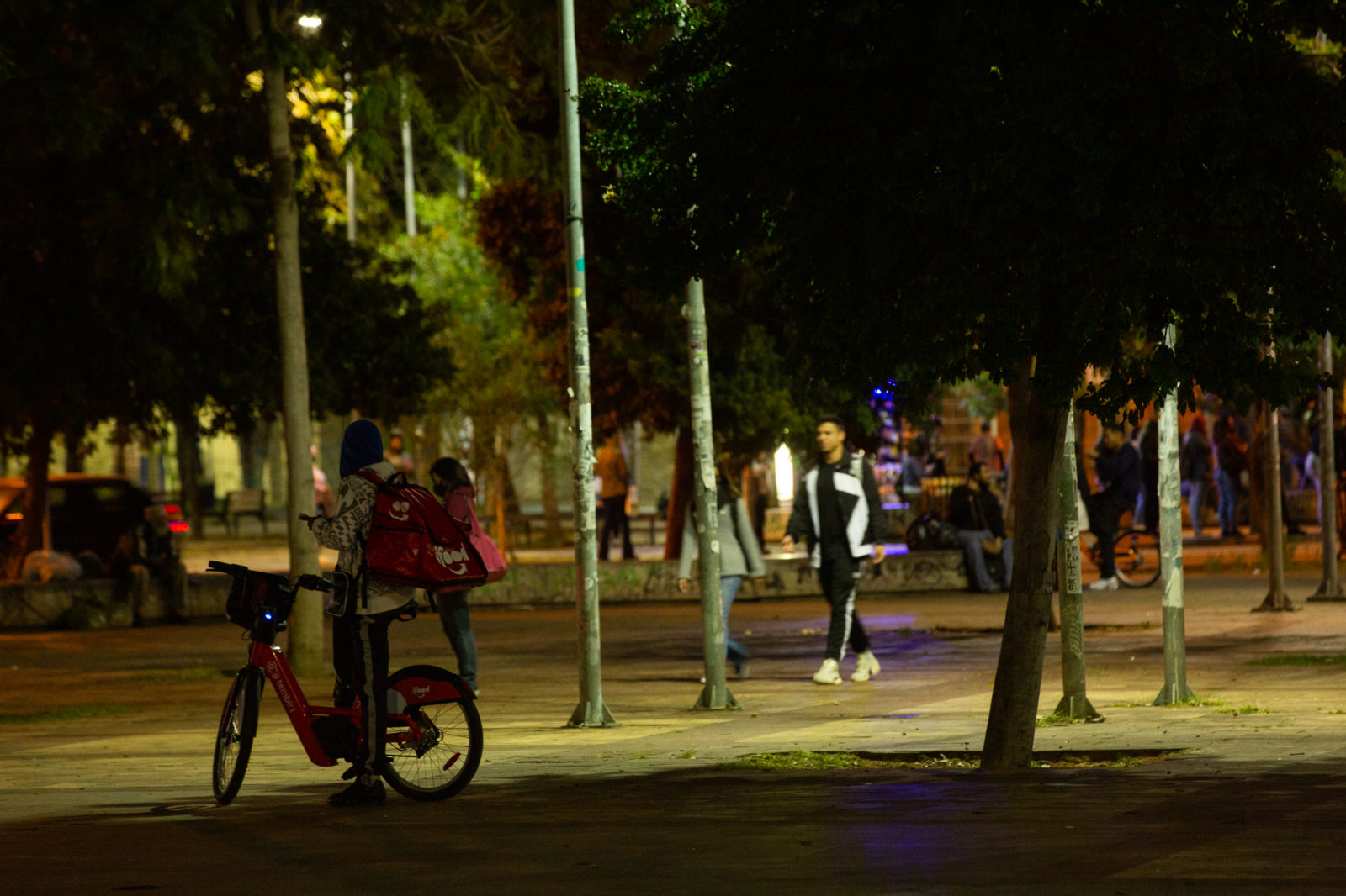 À espera da próxima entrega, uma adolescente trabalha a noite com sua mochila vermelha do Ifood e uma bicicleta