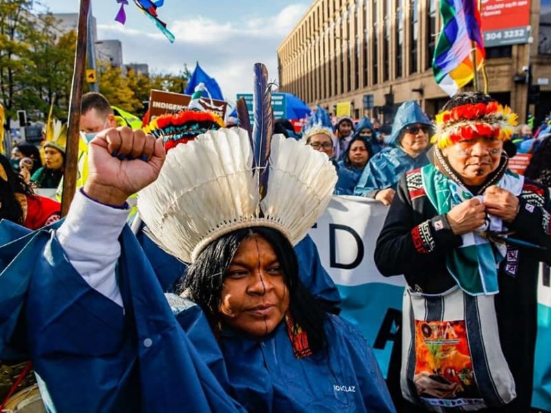 Na foto, Sônia Guajajara em um protesto em Glasgow com outros indígenas ao seu redor. Ela veste adereços indígenas e está com punho cerrado em riste
