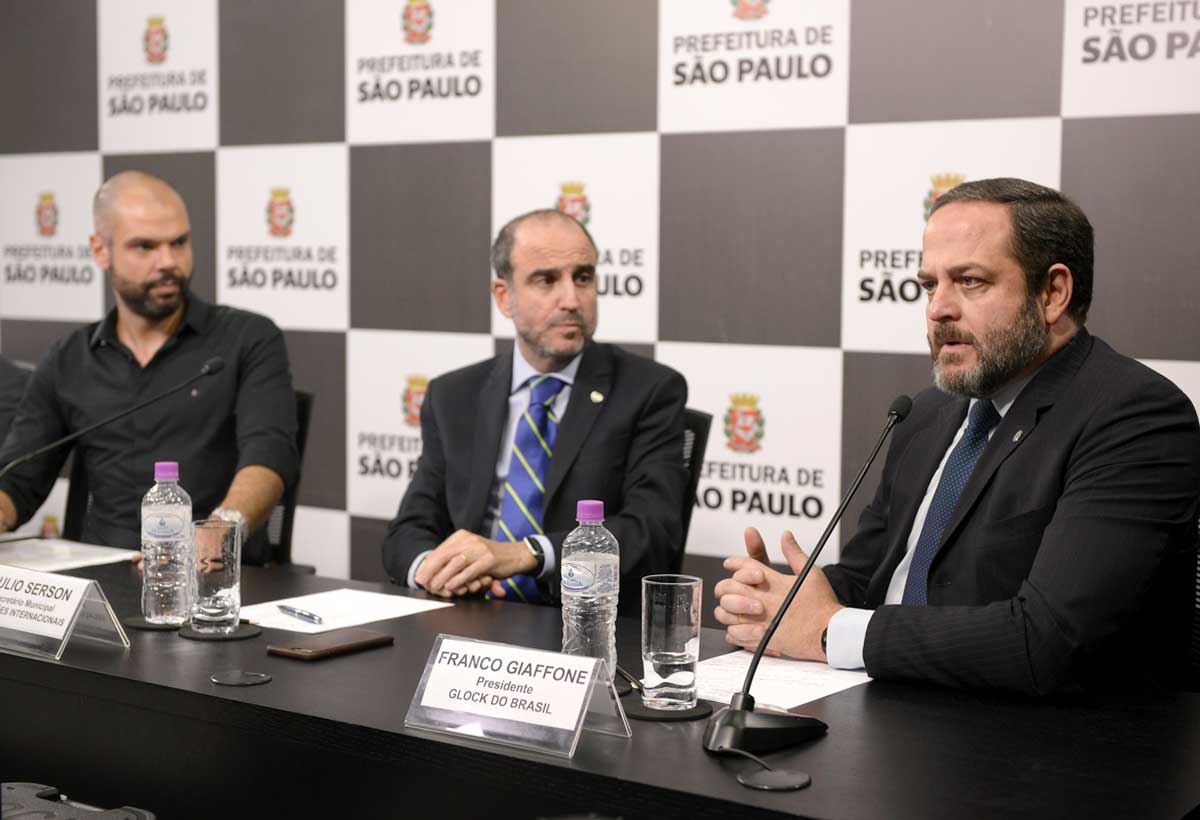 A foto apresenta três homens brancos; à esquerda, Bruno Covas, ex-prefeito de São Paulo e à direita Franco Giaffone, empresário patrocinado pela Glock