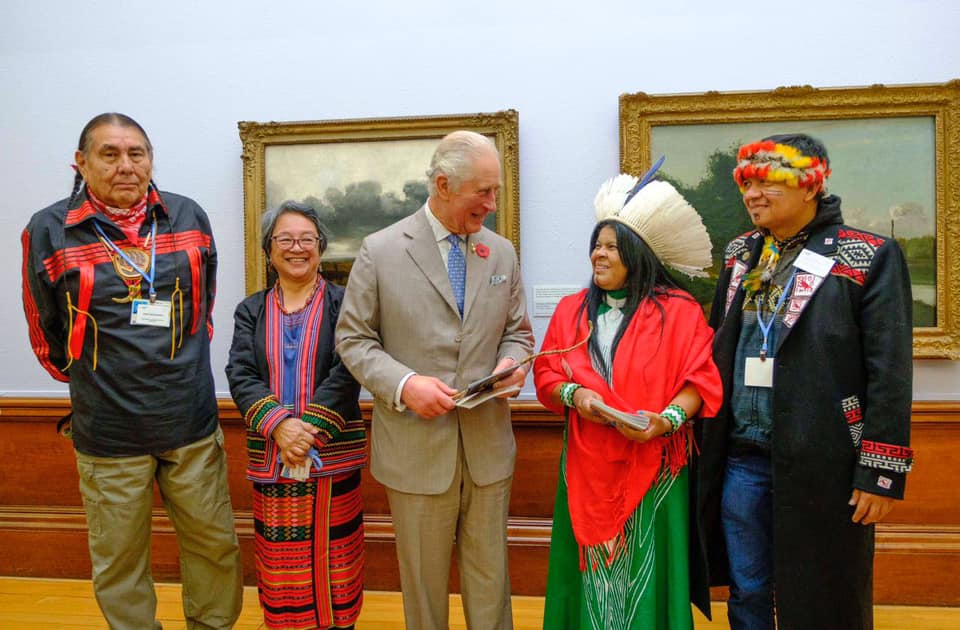 Quatro indígenas posam ao lado do príncipe Charles, um homem branco de terno marrom, durante evento da COP-26. Ambos os indígenas, dois homens e duas mulheres, usam alguns adereços tradicionais