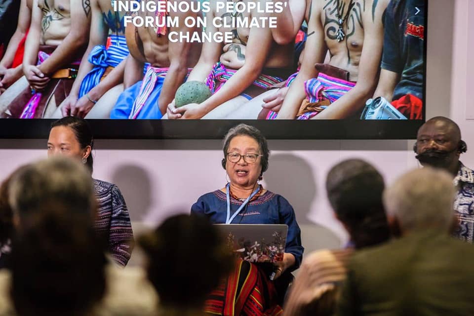 Victoria Tauli é uma mulher indígena asiática, que veste uma blusa azul e óculos. Ao seu lado estão um homem e uma mulher; atrás dela um painel escrito "Fórum de Mudança Climática"