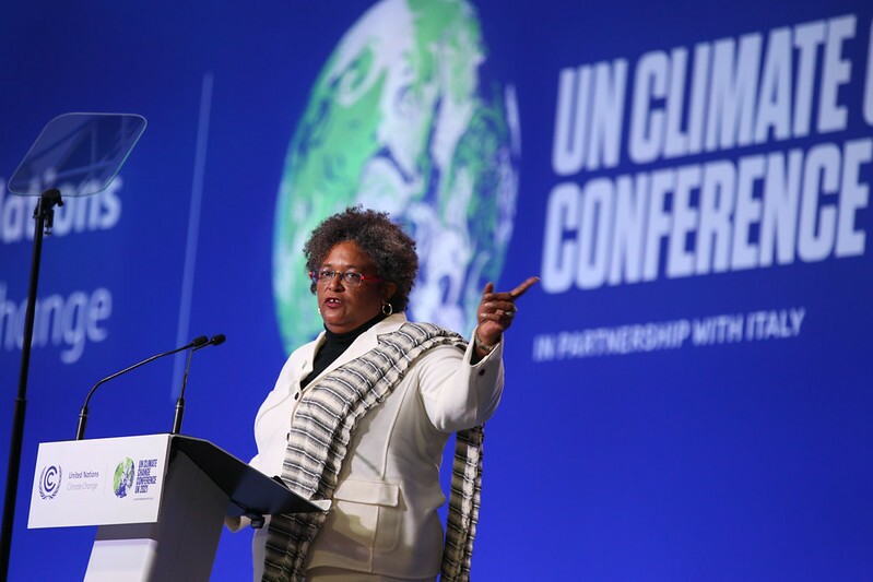 Mia Mottley, uma mulher negra e primeira-ministra de Barbados em discurso na COP-26; ela veste uma roupa branca e posa em frente a um painel azul com o globo terrestre atrás