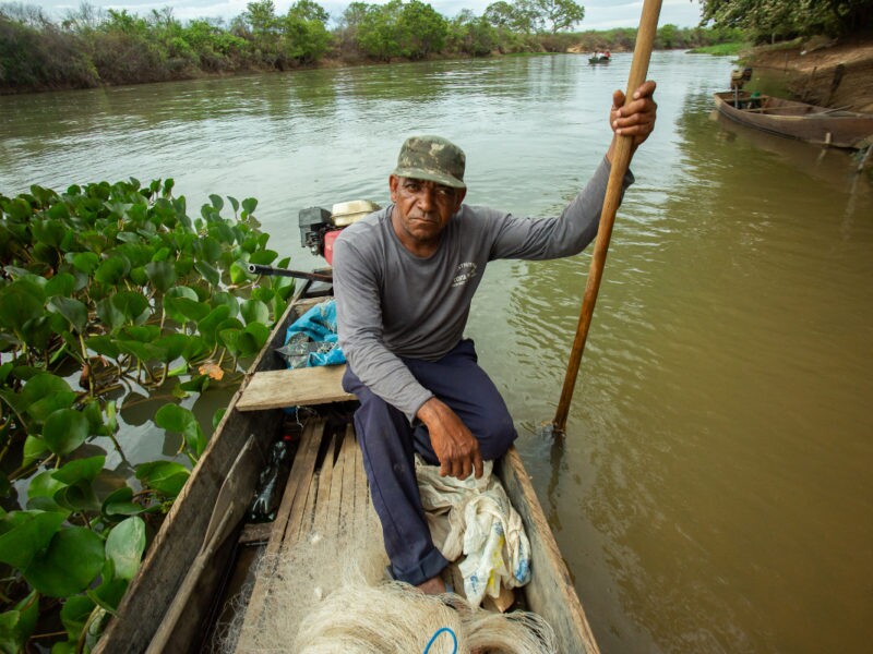 Homem negro posa sentado em um barco à beira de um rio; ele veste calça e blusa de mangra longa cinza, um boné verde escuro e segura na mão direita um bastão de madeira