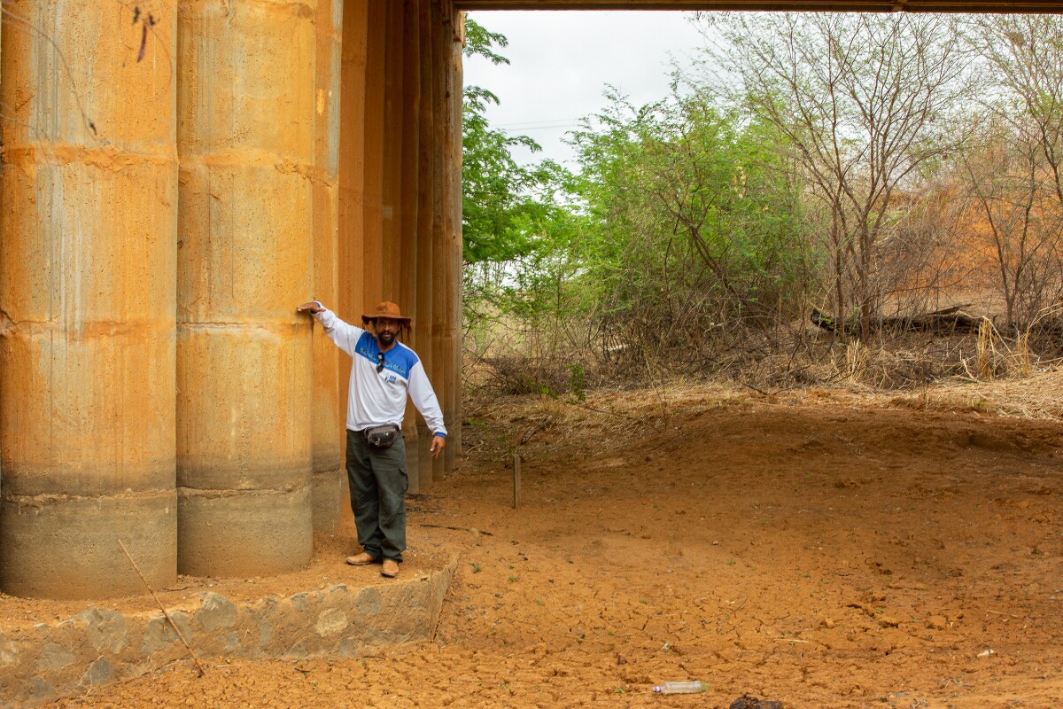 Um homem negro vestindo uma camiseta branca com a azul, chapéu marrom e calça jeans posa em frente a pilastra de uma ponte, mostrando em qual altura o rio costumava correr