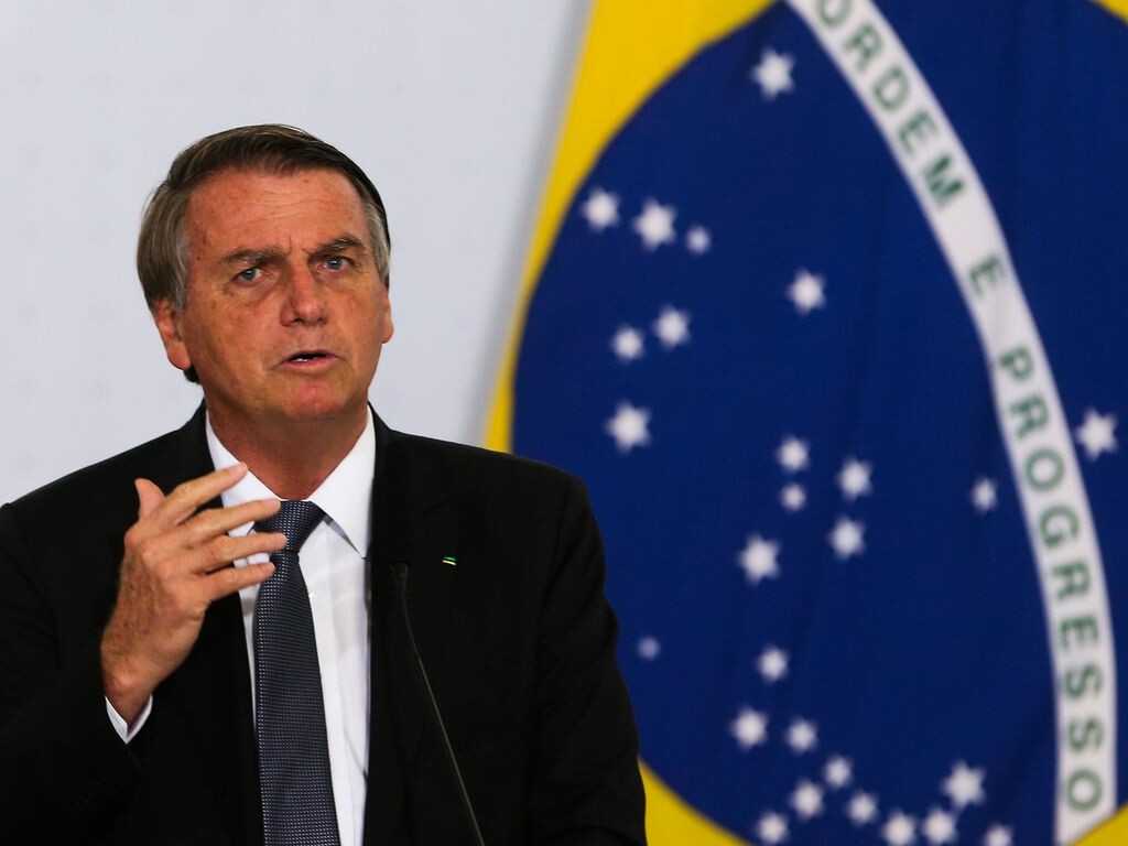 Presidente Bolsonaro em frente à uma bandeira do Brasil; ele veste um terno preto com camisa branca