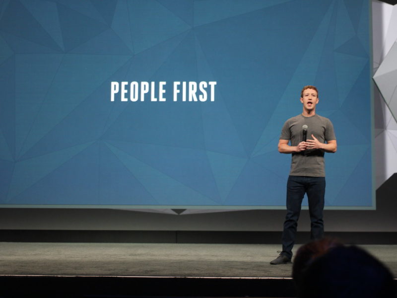 Zuckerberg é um homem branco de cabelos castanhos, ele veste uma calça jeans escura com uma camiseta cinza; atrás dele está um painel azul escrito "people first", ou em tradução livre, "primeiro as pessoas"