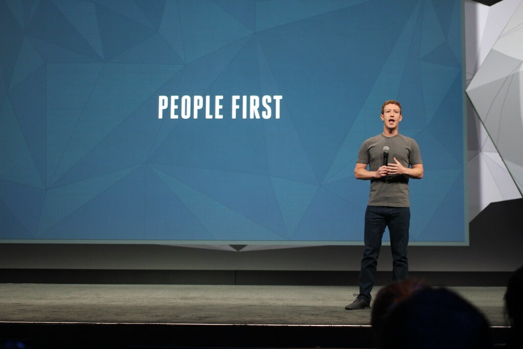 Zuckerberg é um homem branco de cabelos castanhos, ele veste uma calça jeans escura com uma camiseta cinza; atrás dele está um painel azul escrito "people first", ou em tradução livre, "primeiro as pessoas"