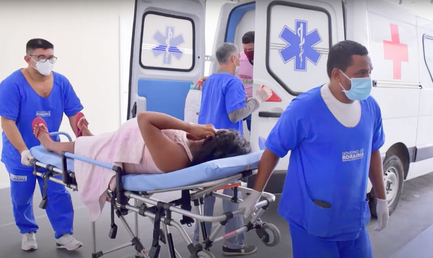 Na foto, uma mulher grávida é transportada de ambulância; ela está deitada em uma maca e é acompanhada por dois profissionais