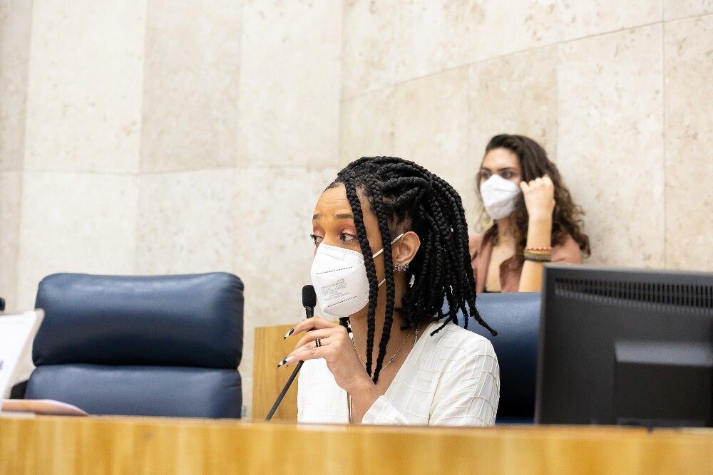 Erika Hilton veste uma camisa e máscara branca; ela está sentada falando ao microfone durante a reunião de instalação da CPI sobre violência contra à população trans