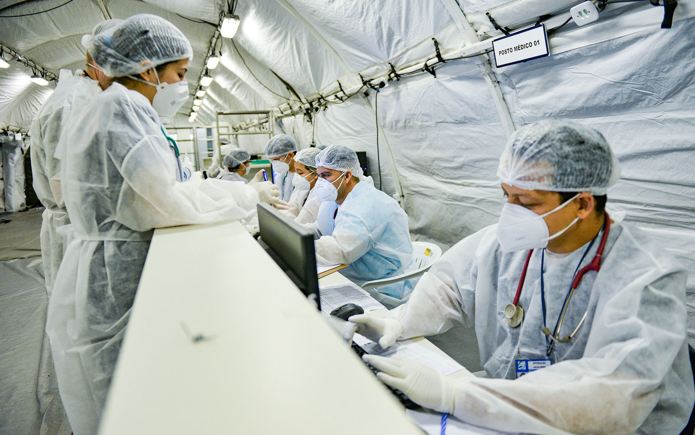 A imagem apresenta médicos do Hospital de Campanha de Roraima sentados em uma mesa, todos vestem equipamentos de proteção individual como jaleco, máscara, touca e luva brancas