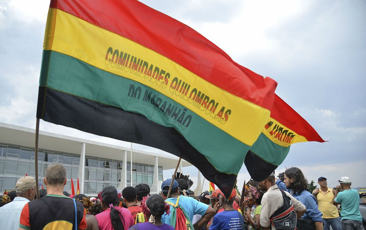 Quilombolas manifestando em frente ao STF; eles seguram uma bandeira nas cores vermelha, amarela, verde e preta com os dizeres 