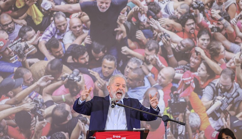 Enquanto gesticula com as mãos, o ex-presidente Lula tem uma imagem sua cercado por pessoas ao fundo