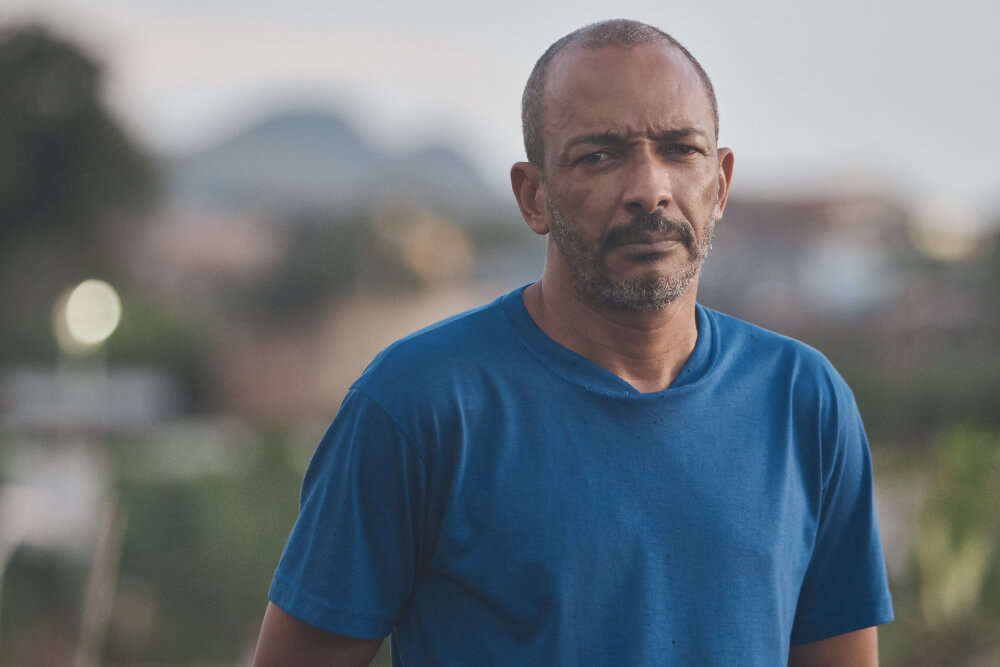 Sobrevivente de Brumadinho, Edivaldo é um homem negro com cabelos grisalhos ralos, ele também tem barba e bigode grisalhos; na foto ele está vestindo uma camiseta azul e o fundo atrás dele está desfocado