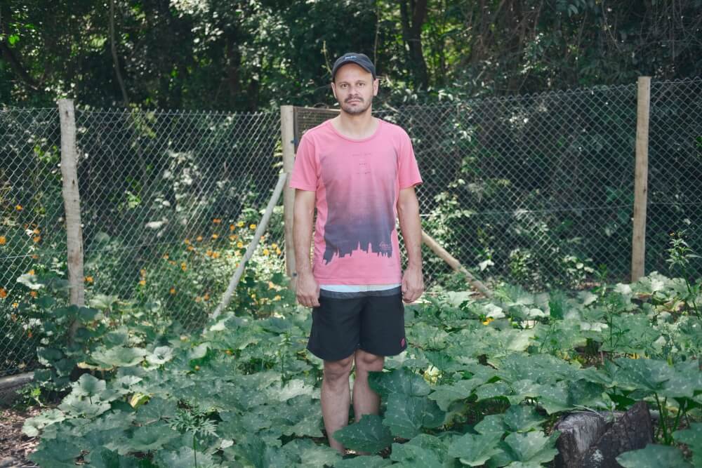 A imagem mostra Ronnie, um homem branco vestindo boné preto, camiseta rosa e shorts preto, parado em meio à sua horta