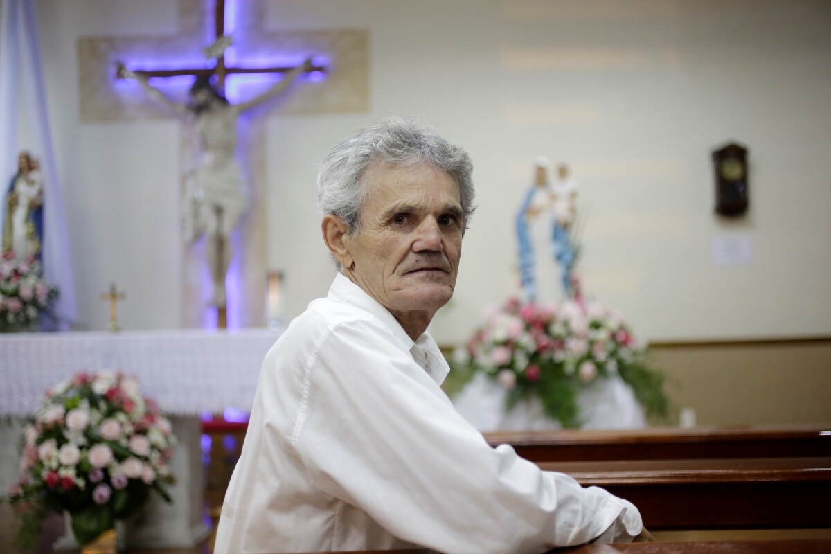 Armandio Seron é um senhor na faixa dos 60 anos, ele é branco com cabelos grisalhos; vestindo uma camisa branca, Armandio está sentado em um banco de uma igreja, atrás dele tem um altar com santos