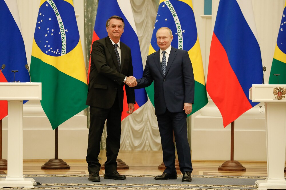 Com as bandeiras do Brasil e da Rússia atrás, Bolsonaro e Putin posam para a imprensa com as mãos apertadas; ambos são homens brancos na faixa dos 60 anos, eles vestem terno preto