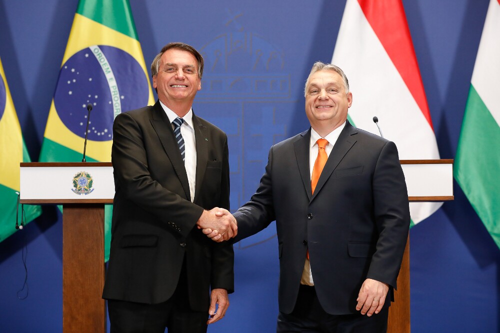 Com as bandeiras do Brasil e da Hungria atrás, Bolsonaro e Orbán posam para a imprensa com as mãos apertadas; ambos são homens brancos na faixa dos 60 anos, eles vestem terno preto