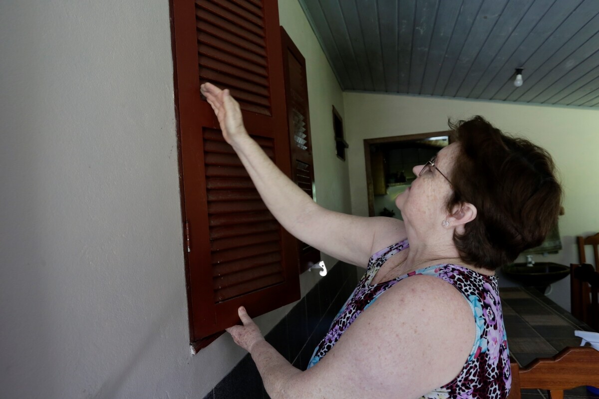 Fátima é uma senhora na faixa dos 60 anos, com cabelos e olhos castanhos; na foto ela está passando as mãos na janela de sua casa e mostrando a poluição da exploração de carvão que acontece em um local próximo
