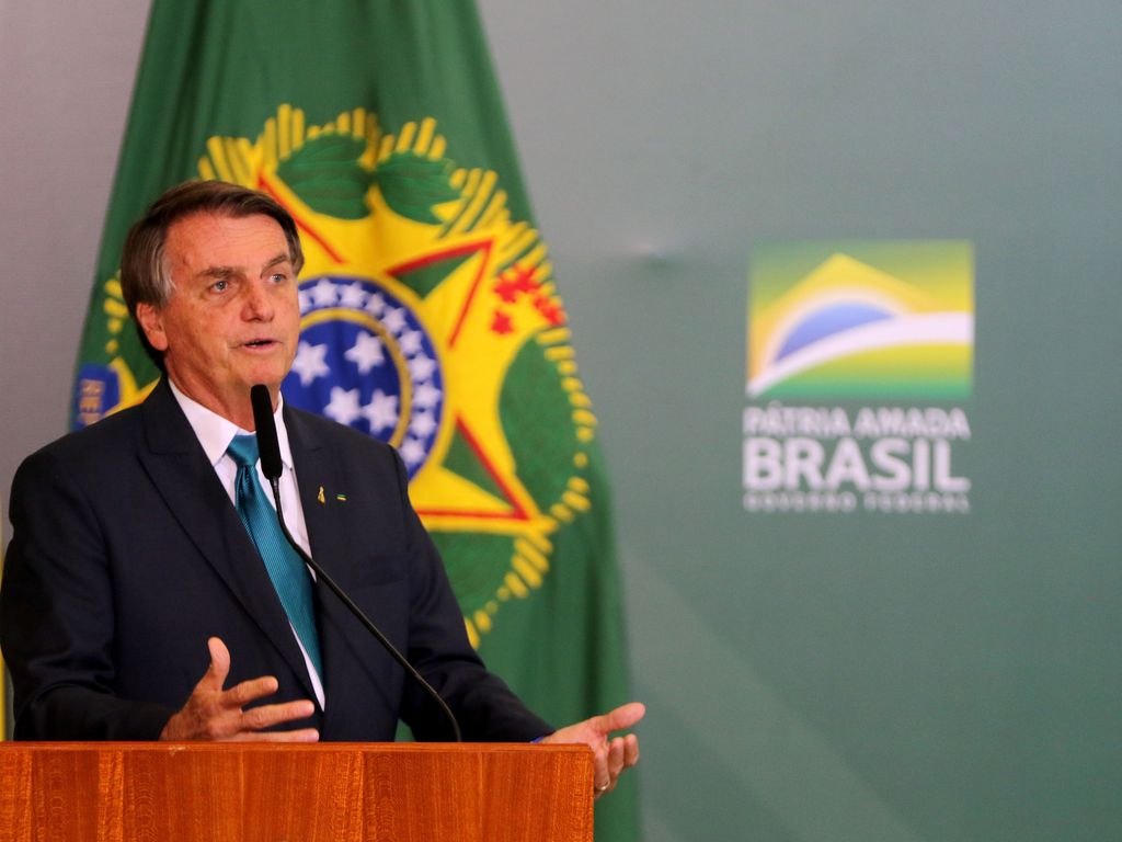 O  Presidente da República Jair Bolsonaro durante evento no Palácio do Planalto; ele veste um terno preto e posa em frente a bandeira do Brasil