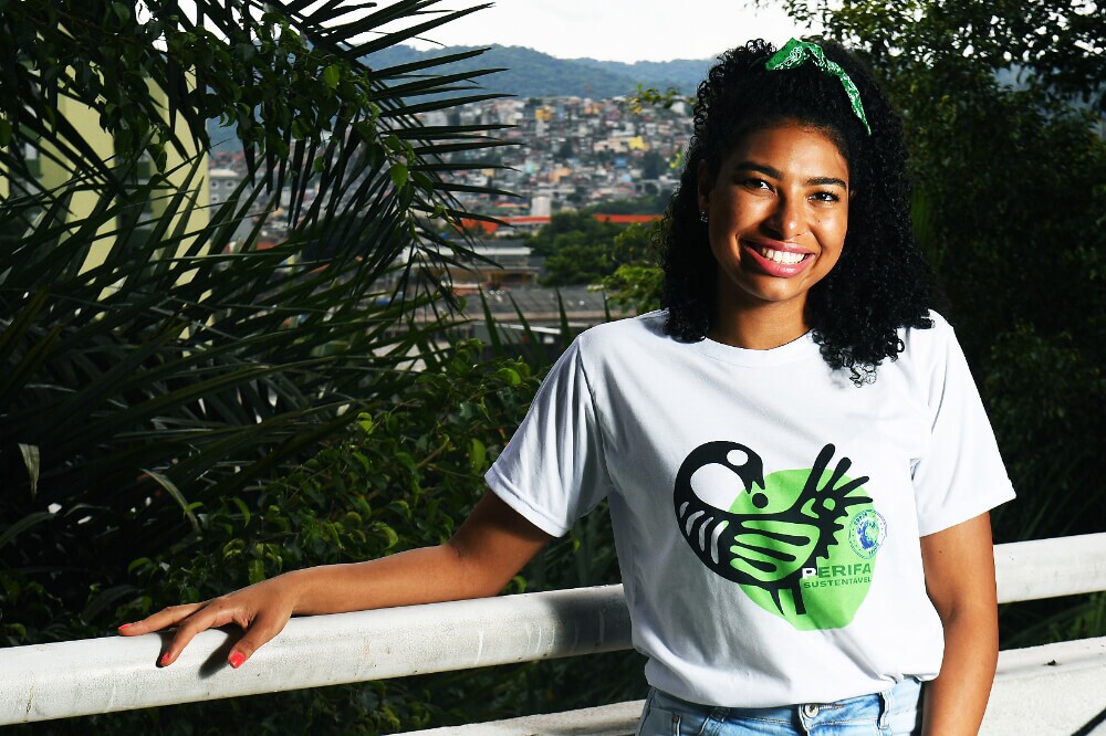 Amanda Costa é uma mulher negra com cabelo cacheado e olhos castanhos; ela veste uma blusa branca com o desenho de um pássaro em preto e os dizeres "periferia sustentável"; atrás dela está a favela onde nasceu, em São Paulo