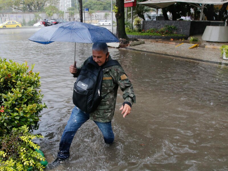 Homem segurando guarda-chuva tenta atravessar rua alagada, com água nos joelhos