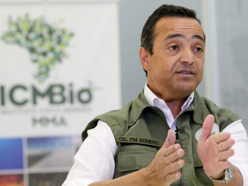 Ex -presidente do ICMBio, Homero George Cerqueira, é um homem branco com cabelos e olhos castanhos; ele veste uma camisa branca com um colete verde militar por cima, atrás dele está um banner do ICMBio, com um mapa verde do Brasil