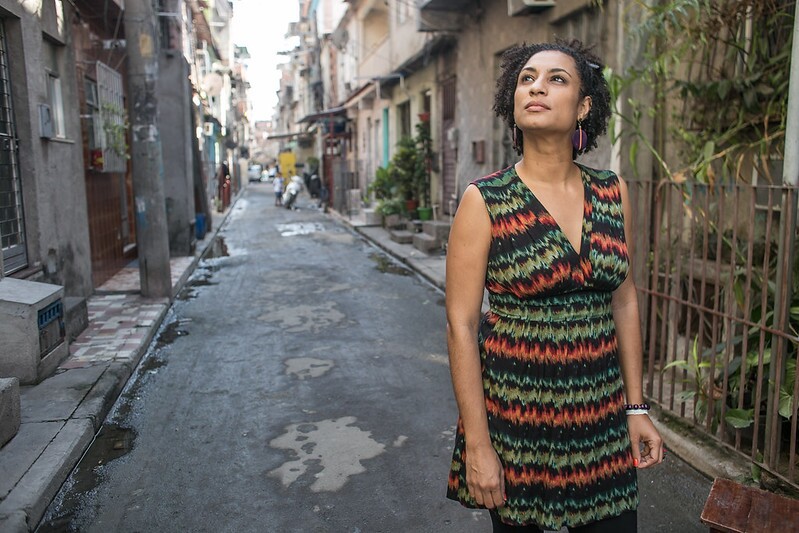 Marielle Franco era uma mulher negra com olhos e cabelos castanhos; na foto ela está usando um vestido colorido, atrás dela está uma rua de uma favela do Rio de Janeiro