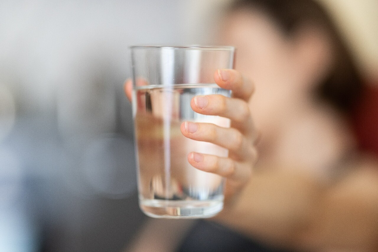 Imagem mostra a mão de uma mulher segurando um copo cheio com água; o fundo da foto está desfocado, sendo visível somente o copo