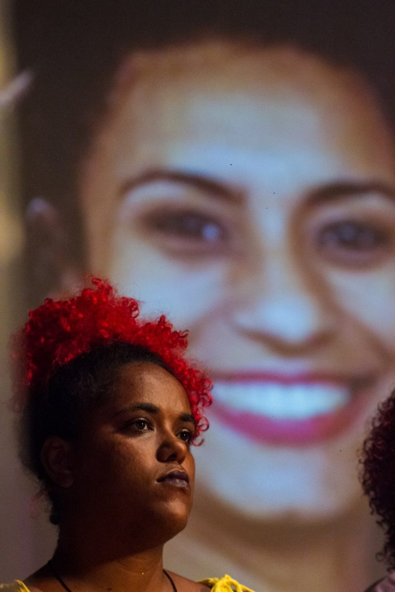 Dani monteiro é uma mulher negra, ela usa um adereço vermelho no cabelo; Ao fundo de Monteiro está uma imagem de Marielle Franco