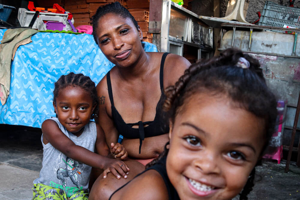 Nina é uma mulher negra com cabelos e olhos castanhos; na foto ela está acompanhada de duas crianças, seus filhos mais novos com  4 e 5 anos