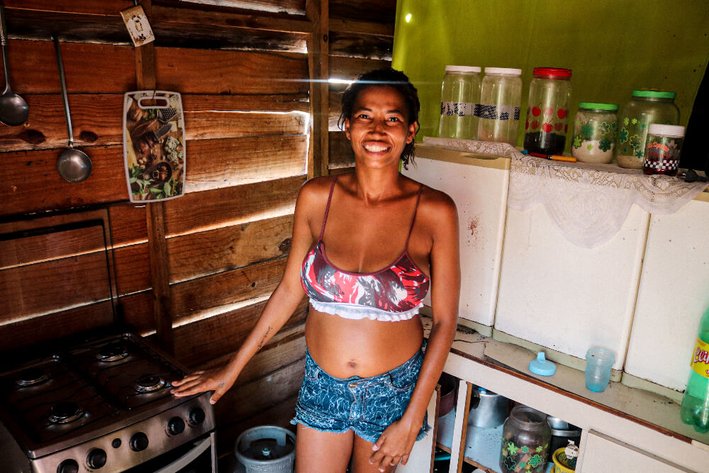 Elaine é uma mulher negra de 32 anos, ela veste um top florido e um shorts jeans; na imagem ela mostra a cozinha de sua casa, um cômodo simples