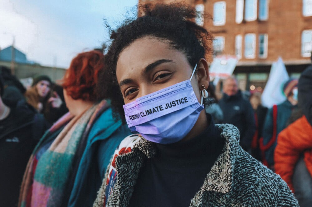Karina Penha é uma mulher negra com cabelos e olhos castanhos; a imagem mostra Karina olhando para a câmera durante uma manifestação, ela usa uma máscara com os dizeres "climate justice"