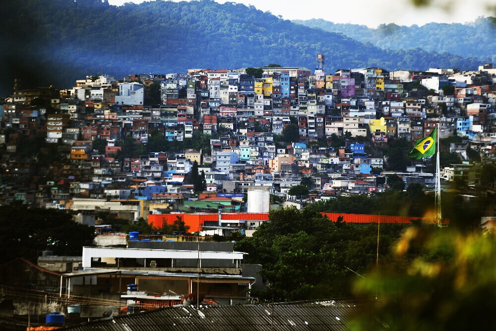 Imagem mostra ao fundo região periférica da cidade de São Paulo, com casas em um morro próximo a uma área verde