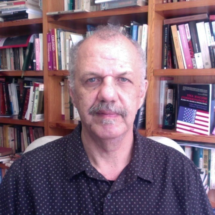 Nasser é um homem na faixa dos 60 anos, branco, com olhos claros e cabelos e bigode grisalhos. Ele veste uma camisa preta de bolinhas e posa em frente a uma estante de livros