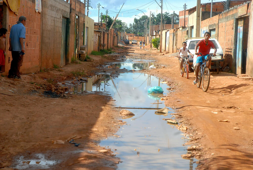 Rua em favela mostra ausência de saneamento básico, com água suja correndo enquanto crianças brincam de bicicleta