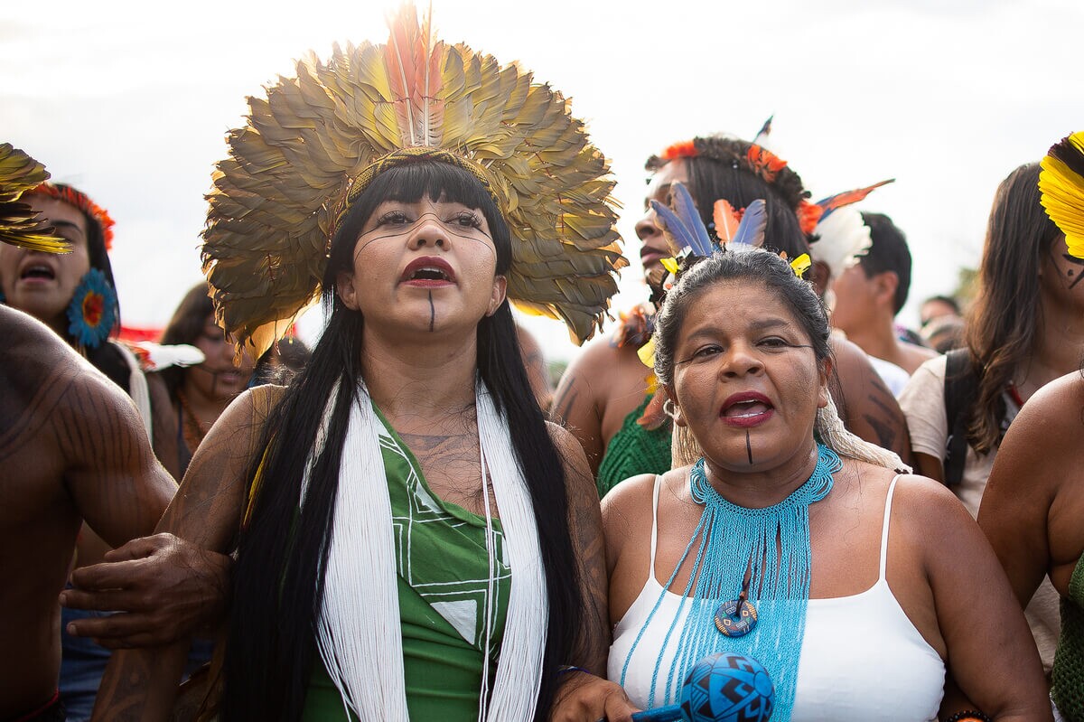 À esquerda da foto está uma mulher indígena com cabelos longos e franja castanho escuro; ela veste uma blusa verde musgo e adereços tradicionais na cabeça e no pescoço, além de pintura no rosto. Ao seu lado está outra mulher indígena, com cabelo castanho preso em um coque; ela veste regate branca e adereço tradicional no cabelo e pescoço.