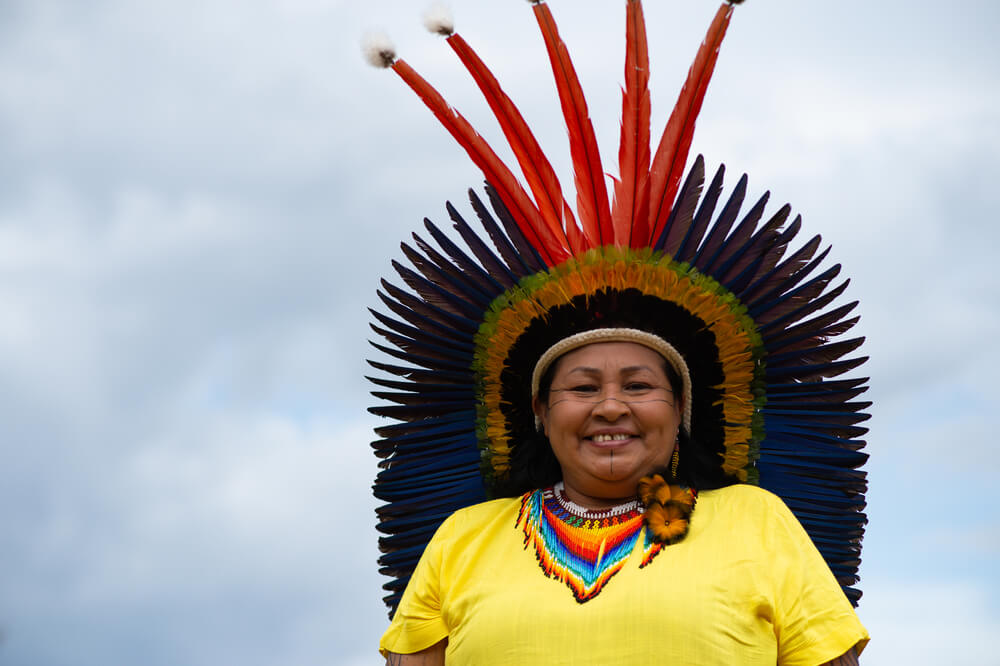 Telma é uma mulher indígena com olhos e cabelos castanhos; ela usa um cocar na cabeça e uma camiseta amarela