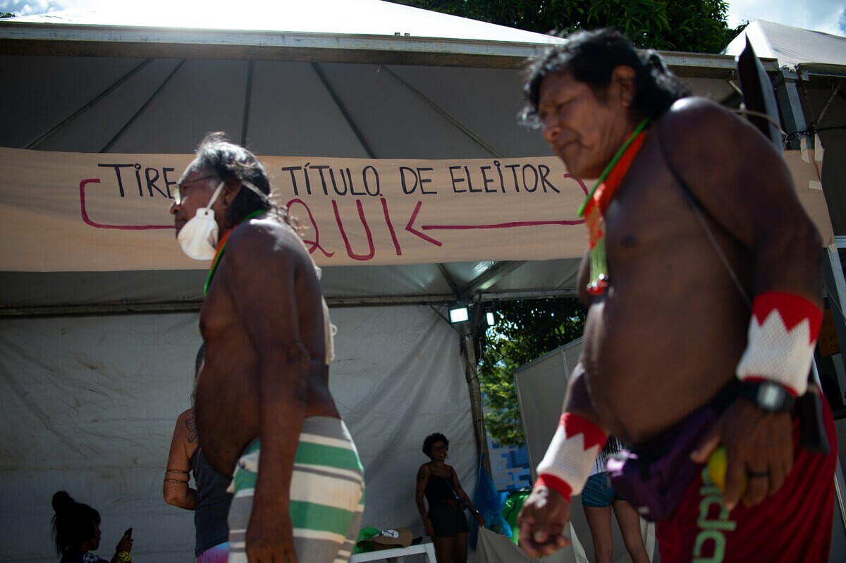 Barraca montada em acampamento indígena com cartaz pendurado na entrada com os dizeres "tire seu título de eleitor aqui"; dois homens indígenas passam em frente à barraca