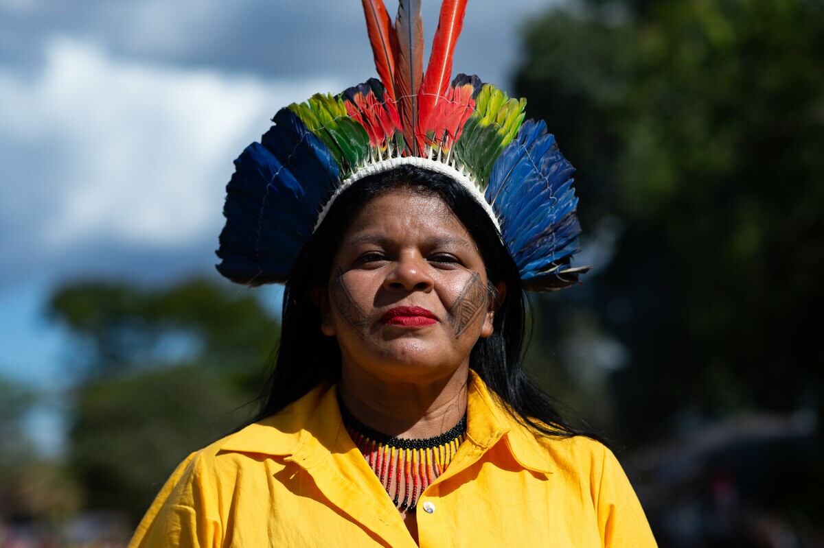 Mulher indígena olha diretamente para a foto; ela veste camisa amarela vibrante e um adereço indígena no pescoço e na cabeça; além disso, usa batom vermelho e pintura facial