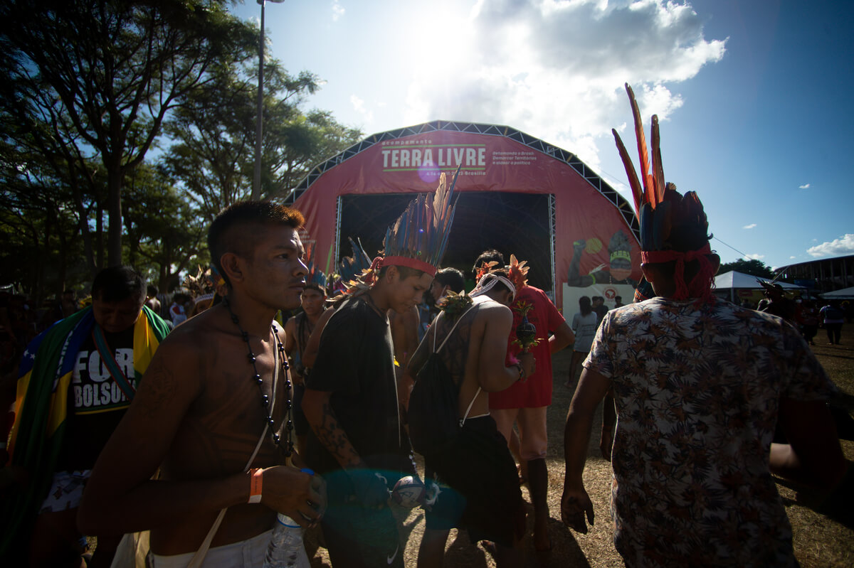 Homens indígenas se reúnem em frente a palco do acampamento Terra Livre; eles usam adereços tradicionais