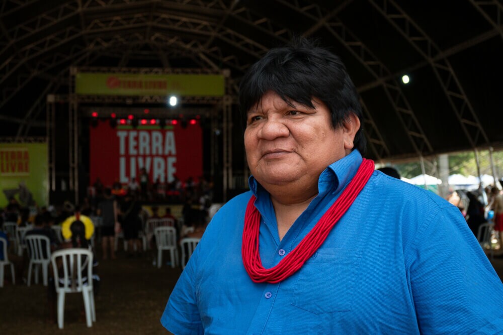 Almir é um homem indígena com cabelos curtos e escuros; ele usa uma camiseta azul e um colar tradicional vermelho
