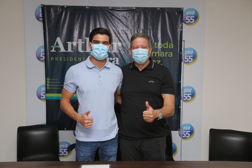 João Catunda, à esquerda, é um homem branco na faixa dos 20 a 30 anos, com cabelos castanhos; ele veste máscara descartável azul e camiseta polo azul. Ao seu lado está Arthur Lira, um homem branco na faixa dos 40 anos, com cabelos grisalhos e olhos castanhos; ele usa máscara descartável azul e uma camiseta polo preta