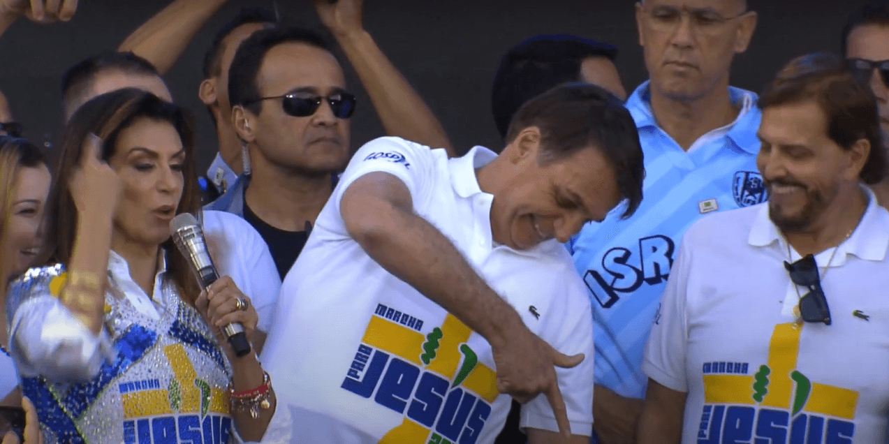 Presidente Bolsonaro usa camiseta branca com o logo da Marcha Para Jesus e posa ao lado de pastores fazendo arminha com a mão